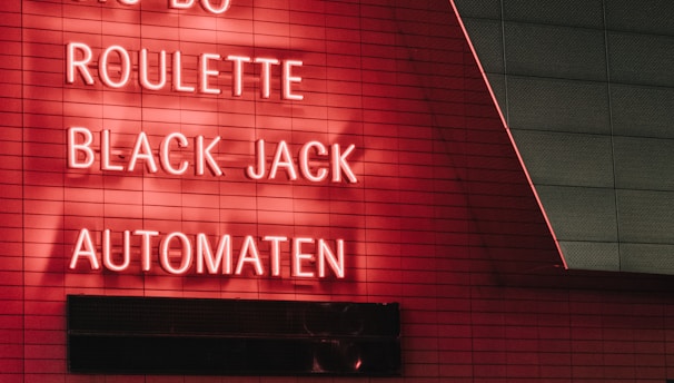 Red neon letters are mounted on a dark tiled wall, displaying the words Poker, Sic Bo, Roulette, Black Jack, and Automaten.